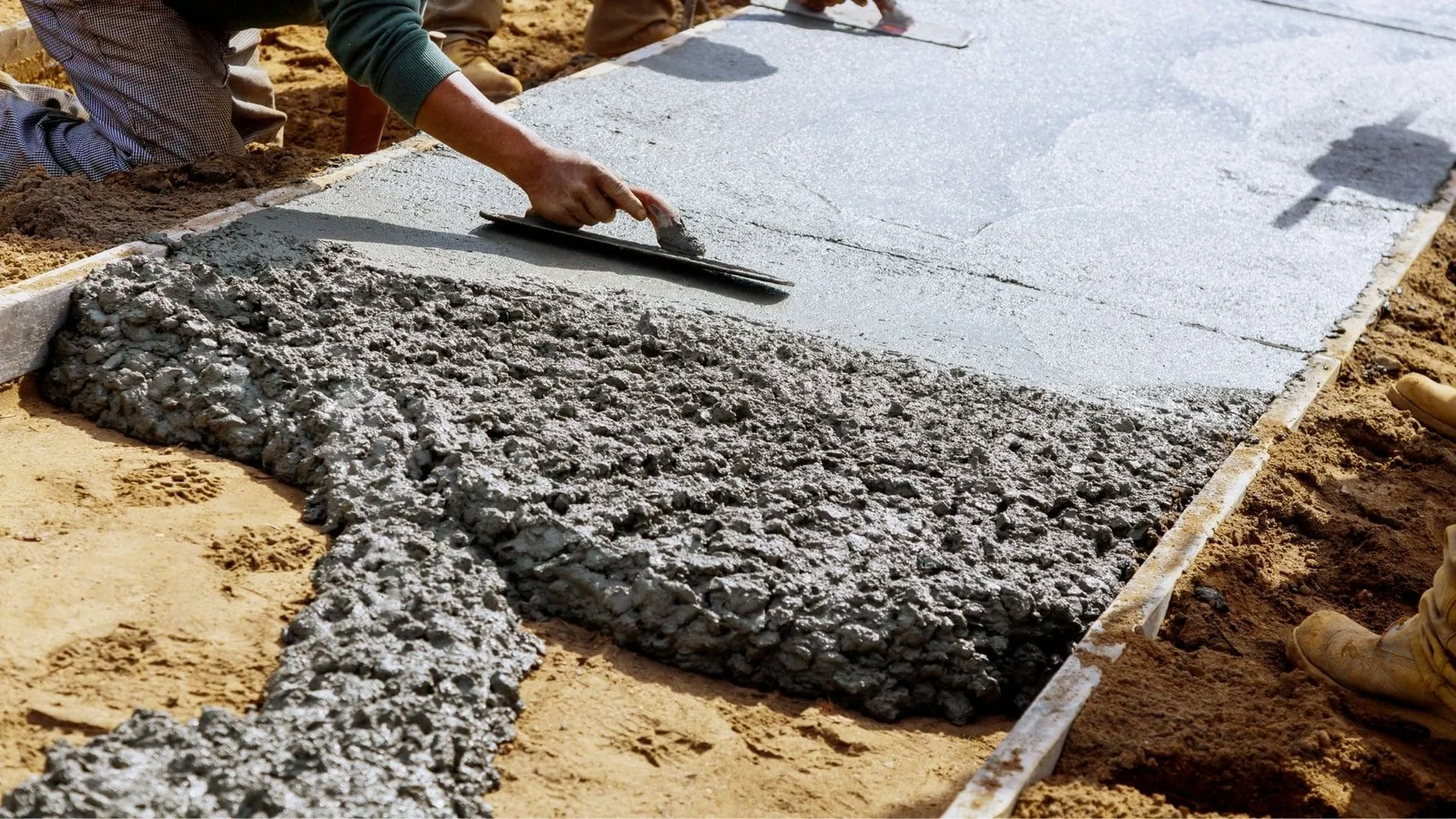 Worker finishing fresh concrete slab with trowel, wooden forms visible along edges